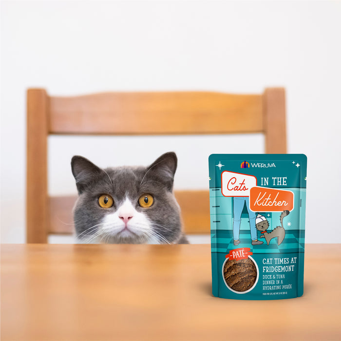 A gray and white cat sits behind a wooden table, staring curiously at a packet of Cats in the Kitchen Cat to The Future cat food placed in front of it.
