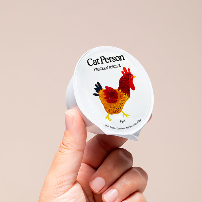 A hand holds a small container of Cat Person Chicken Paté, with a chicken illustration on the lid. This protein-rich cat food offers balanced nutrition, shown against a plain beige background.