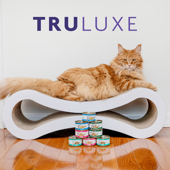 A fluffy orange cat lounges on a modern white scratcher with several cans of TruLuxe Kawa Booty wild-caught tuna cat food stacked in front. The word TruLuxe appears in large purple letters above the cat.